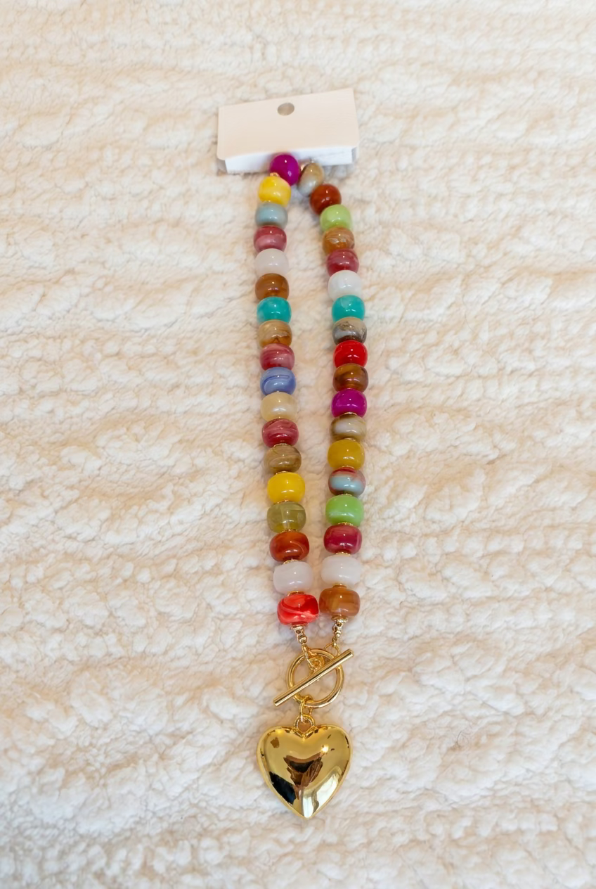 A colorful beaded necklace with a gold heart-shaped pendant, laid flat on a textured cream fleece blanket. The beads feature multiple colors including red, yellow, green, blue, and purple, with a small white paper tag attached at the top.