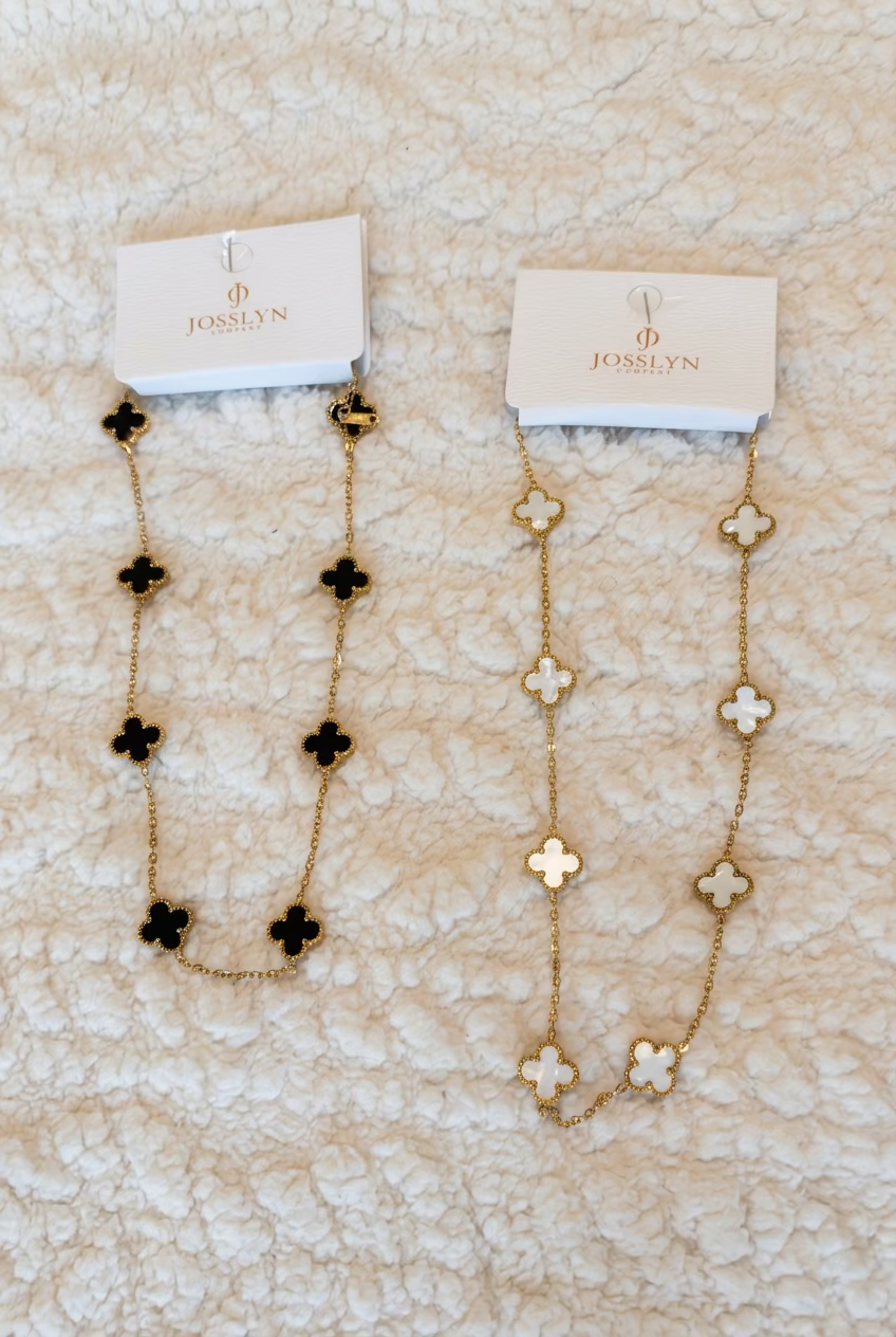 Two gold-tone chain necklaces with clover-shaped charms laid flat on a cream sherpa fabric background. The top necklace has white inlay clovers, and the bottom necklace has black inlay clovers.